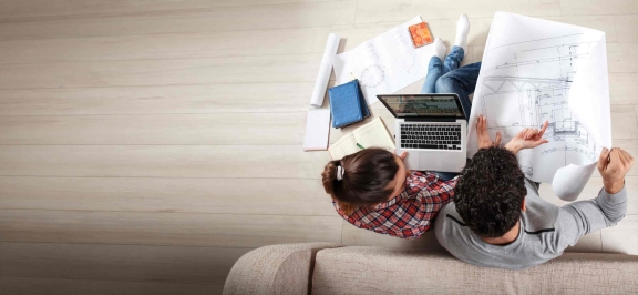 Frau und Mann sitzen mit Laptop und Bauplan beisammen. Man sieht die beiden aus der Vogelperspektive.